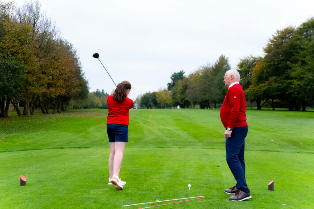 Golfer swings on a lush golf course with trees in the background, wearing a red top and blue shorts.