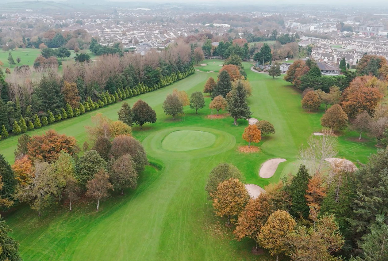 Aerial view of a lush green golf course with autumn trees, sand bunkers, and a suburban backdrop.